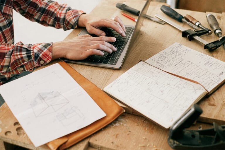 A person working on their laptop in a workshop, with tools and notepads on their bench